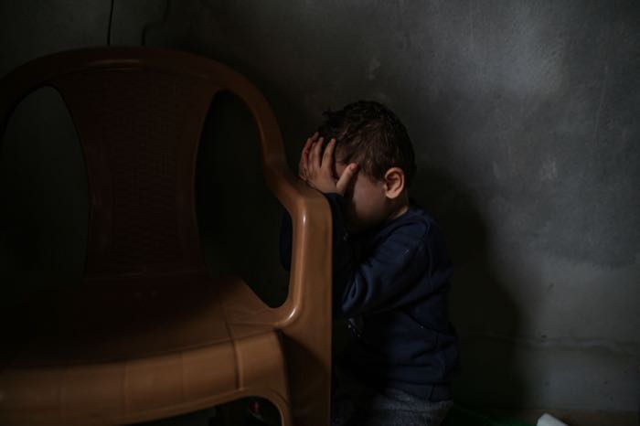 A boy sits alone against a wall, covering his face.