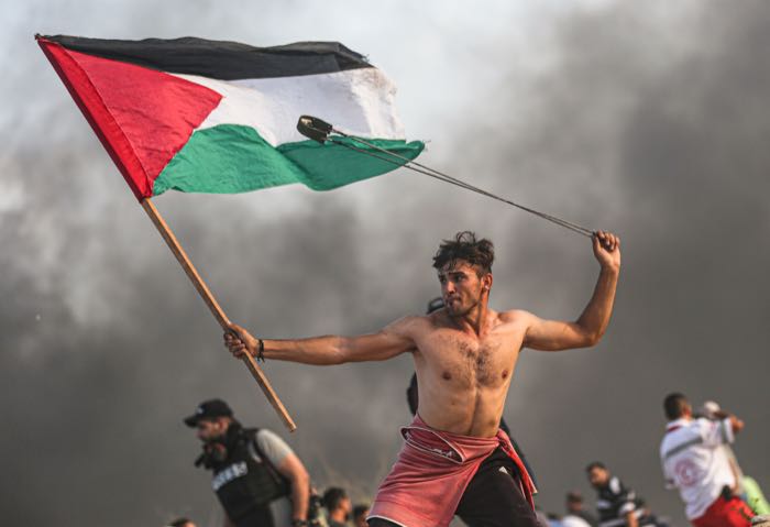 A young man carries a flag and a slingshot through smoke.