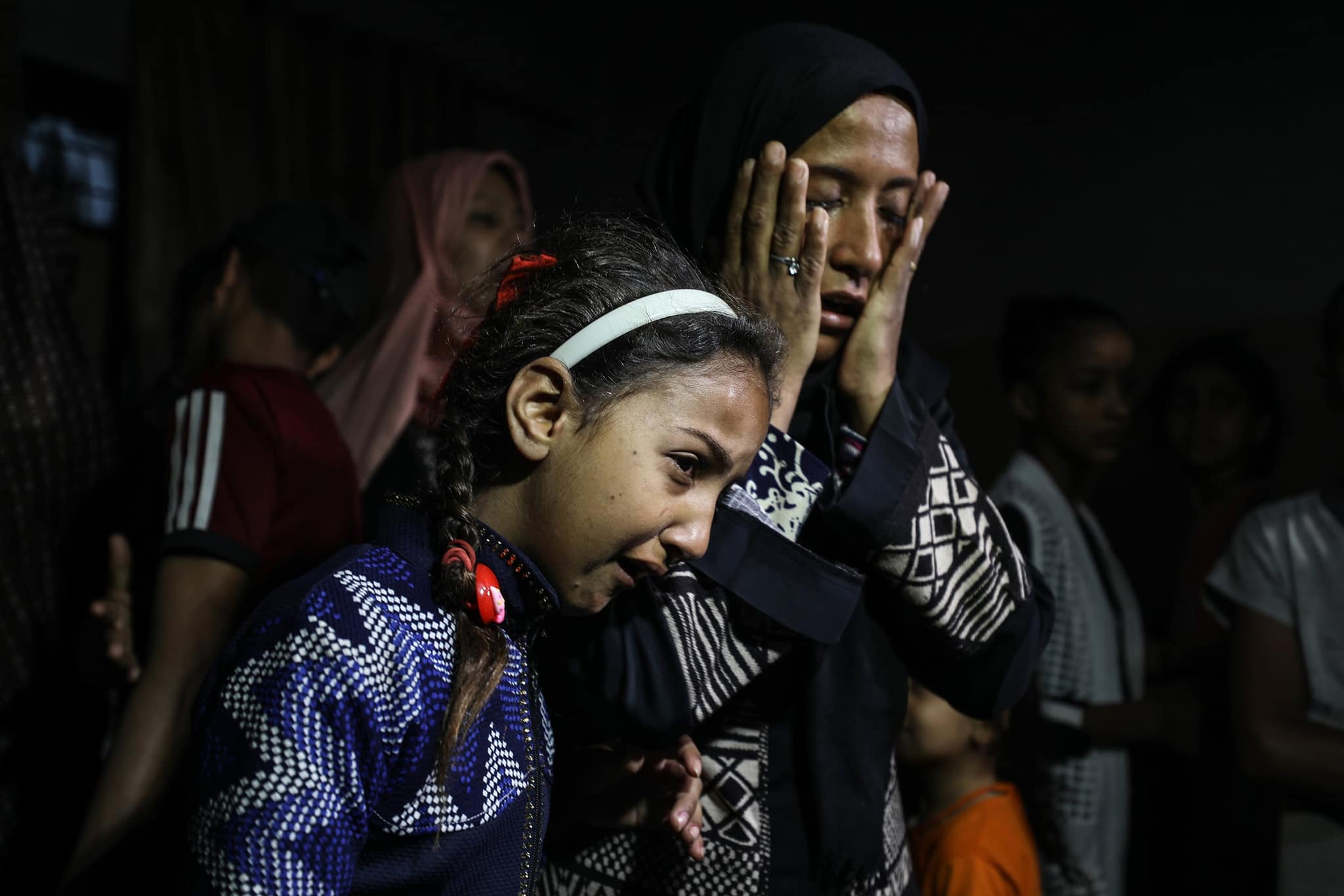 امرأة تمسح دموعها وفتاة تستند إليها في غرفة معتمة.