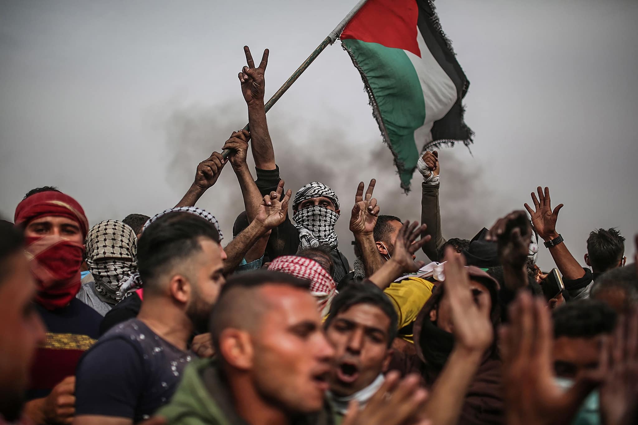 متظاهرون يرفعون علمًا وإشارات النصر أمام سماء مثقلة بالدخان.